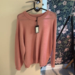 Rosa stickad tröja från 157 - Mysig rosa stickad tröja från 157 med rund halsringning och långa ärmar. Enkel och stilren design som passar perfekt till jeans eller kjol. Tröjan har ribbade muddar vid ärmslut och nederkant för en snygg finish.