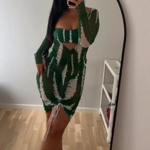 3 delat set - Säljer en grön och vit kortklänning med coolt randigt mönster. Klänningen har lång ärm, omlottdetalj framtill och snörning nertill. Tight passform och öppen detalj vid magen gör den extra snygg. Perfekt för dig som vill sticka ut! Samt bikini och trosa till