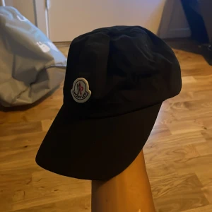 Svart keps från Moncler - Snygg svart keps från Moncler med klassisk böjd skärm och broderad logotyp framtill. Kepsen har en stilren design och är tillverkad i ett lätt och bekvämt material som passar perfekt till streetwear eller sportiga outfits.