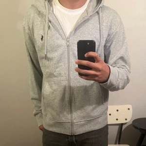Grå hoodie från Polo Ralph Lauren - Snygg ljusgrå hoodie från Polo Ralph Lauren med dragkedja och klassisk broderad logga på bröstet. Tröjan har huva med snörning, kängurufickor och är tillverkad i mjuk bomullsmix. Perfekt för chill dagar eller när du vill ha en clean streetstyle.