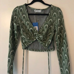 Grön mönstrad croppad topp Bershka - Säljer en grön långärmad croppad topp från Bershka med coolt grafiskt mönster. Toppen har v-ringning, knytning framtill och långa band som hänger ner. Perfekt för dig som gillar trendiga detaljer och snygg passform.