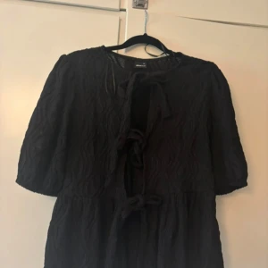 Svart omlott topp från Gina Tricot - Svart topp från Gina Tricot med knyt framtill och lätt struktur i tyget. Toppen har halvlånga ärmar och en avslappnad passform. Perfekt för dig som gillar en stilren look med lite edge.