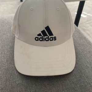 Vit keps från Adidas med svart logga - Snygg vit keps från Adidas med klassisk svart logga framtill. Justerbar rem baktill för perfekt passform. Kepsen har böjd skärm och ventilerande hål på ovansidan. Perfekt accessoar för en sportig och clean look.