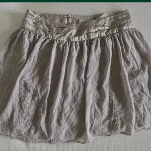 Beige kortkjol från Zadig & Voltaire - Supersöt beige kortkjol från Zadig & Voltaire med broderad detalj nertill. Kjolen har ett brett, glansigt pals midjeband med knappar framtill och är gjord i ett tunt, luftigt tyg som ger en avslappnad vibe. Perfekt för dig som gillar en feminin och chill stil. Köpt i Paris. Färg ljus lila 