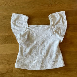 Vit blus med broderade detaljer Kappahl - Superfin vit blus från Kappahl med broderade hål och puffiga korta ärmar. Toppen har fyrkantig ringning och är gjord i ett lätt bomullstyg som känns luftigt och fräscht. Perfekt för en somrig look med extra söt vibe.