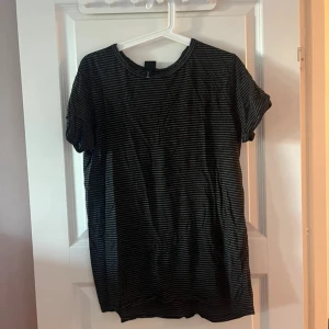 Svart och grå randig t-shirt - En svart t-shirt med tunna grå ränder och rund hals. Modellen är klassisk med korta ärmar och en avslappnad passform. Perfekt för en chill vardagslook och enkel att matcha med jeans eller shorts.