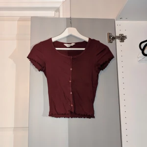 Vinröd croppad topp med knappar - Supersöt vinröd croppad topp med korta ärmar och knappar framtill. Toppen har ribbad struktur och volangkant vid ärmslut och nederkant. Perfekt att styla med jeans eller kjol för en trendig look.