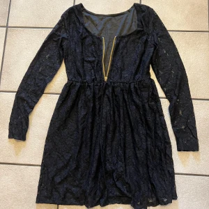 Svart spetsklänning med dragkedja storlek S/M - Svart kortklänning i spets med långärmade ärmar och en snygg guldig dragkedja framtill. Klänningen har markerad midja och ett feminint, lätt utställt kjolparti. Perfekt för dig som gillar detaljer och vill sticka ut lite extra.