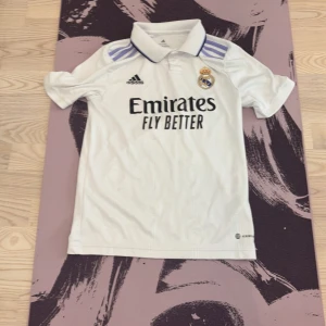 Real Madrid hemmatröja Adidas 11-12 år - Snygg vit Real Madrid hemmatröja från Adidas med klassisk krage och lila ränder på axlarna. Tröjan har klubbmärke och Adidas-logga på bröstet samt trycket 'Emirates Fly Better' framtill. Tillverkad i lätt och ventilerande material.