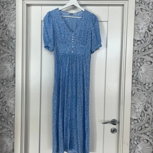 Zudora klänning Indiska - Supersöt långklänning från Indiska, Zudora, i blått med små vita blommor. Klänningen har v-ringning, knappar framtill, korta ärmar och smock i ryggen för extra passform. Perfekt för sommarens alla tillfällen!
