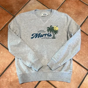 Grå sweatshirt från Morris - Snygg grå sweatshirt från Morris med tryck av palmer, sol och texten 'Hôtel Pelicano' på bröstet. Tröjan har rund hals, långa ärmar och ribbade muddar. Perfekt för en avslappnad och cool stil.