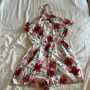 Blommig kort sommarjumpsuit - Supersöt kort jumpsuit med blommigt mönster i rött, rosa och grönt på vit botten. Klänningen har offshoulder-design med smala axelband och resår i midjan för en skön passform. Perfekt för varma dagar och ger en fräsch vibe.