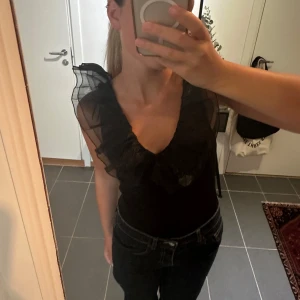 Svart volang body - Säljer en svart body med djup v-ringning och stora volanger längs urringningen och axlarna. Axlarna är gjorda i transparent tyg som ger en cool och unik look. Perfekt för dig som vill sticka ut lite extra! Passar snyggt till både jeans och kjol.