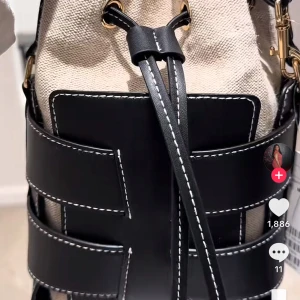 Beige och svart bucket bag med snörning - Snygg bucket bag i beige canvas med svarta detaljer i skinn och vita kontrastsömmar. Väskan har snörning upptill, guldfärgade metalldetaljer och ett coolt rutigt mönster av skinnremmar runt om. Perfekt storlek för det viktigaste och riktigt trendig look. Bor i Gbg så kan mötas och lämna den om ni vill/majorna området men kan åka in till stan osv 