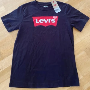 Svart Levi's t-shirt med röd logga - Svart t-shirt från Levi's med klassisk röd logga framtill. T-shirten är kortärmad och har en rund halsringning. Tillverkad i mjuk bomull som känns skön mot huden. Perfekt för en avslappnad och stilren look.