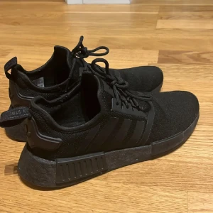 Svarta Adidas NMD sneakers - Säljer ett par helsvarta Adidas NMD sneakers med klassiska tre ränder på sidan. Skorna har stickad ovandel, snörning och en chunky sula med detaljer. Perfekta för dig som gillar en clean och sportig look. 