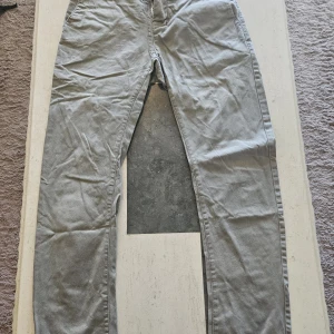 Grå chinos från Wellful, medium - Snygga grå chinos från Wellful i storlek medium. Byxorna har rak passform och är tillverkade i mjukt bomullsmaterial, perfekta för en clean och avslappnad look. Klassisk design med fickor och bälteshällor.