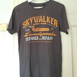 Star Wars vintage look t-shirt  - Cool vintage mörk brun/svart t-shirt från Star Wars med stort tryck på bröstet: 'Skywalker and Son Landspeeder Service and Repair'. T-shirten har rund hals, korta ärmar och är gjord i mjuk bomull. Perfekt för dig som älskar Star Wars och vill sticka ut med en unik tee.