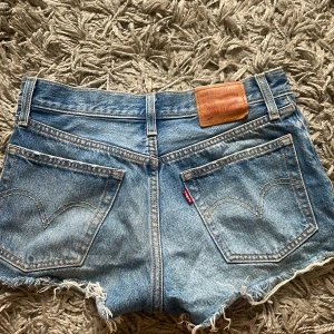 Levi's blå jeansshorts med fransar - Säljer ett par klassiska blå jeansshorts från Levi's med råa fransiga kanter. Shortsen har fem fickor, synliga nitar och den ikoniska Levi's-lappen bak. Perfekta för sommaren och ger en avslappnad vibe. Storlek står ej men midjemått rakt över är 35cm