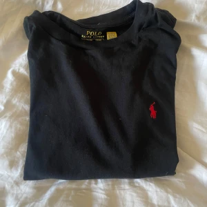 Svart t-shirt från Polo Ralph Lauren - Svart Ralph Lauren T-shirt - Storlek M Snygg och stilren svart T-shirt från Polo Ralph Lauren med klassisk röd logga på bröstet. Passar perfekt både till vardags och festligare tillfällen. Mjuk bomullskvalitet och bekväm passform. Storlek: M Skick: Bra använt skick – inga hål eller fläckar.