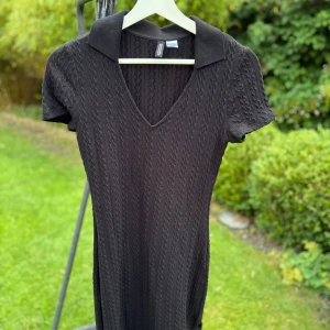 Svart stickad kortklänning H&M Divided - Svart stickad kortklänning från H&M Divided med kabelstickat mönster, v-ringning och krage. Klänningen har korta ärmar och en slim passform som sitter snyggt. Perfekt för dig som gillar en enkel men stilren look.