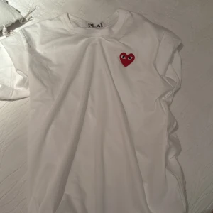 Vit Comme des Garçons Play t-shirt - Vit t-shirt från Comme des Garçons Play med det ikoniska röda hjärtat med ögon broderat på bröstet. Klassisk passform, rund hals och korta ärmar. Tillverkad i mjuk bomull som känns skön mot huden. Perfekt för en clean och stilren look. Den e ganska tajt så den passar Small men storlek M