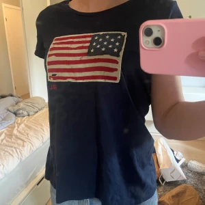 Mörkblå t-shirt från Ralph Lauren - Snygg mörkblå t-shirt från Ralph Lauren med amerikansk flagga broderad på bröstet. Klassisk passform och rund halsringning. Perfekt till jeans eller shorts för en chill look. Hålet i flaggan är design inte slitage! Storleken är XL i barnstorlek, det motsvarar S i vuxen.