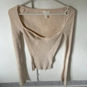 Beige ribbad långärmad topp H&M - Säljer en beige ribbad långärmad topp från H&M med snygg v-ringning och figurnära passform. Toppen har stretchigt material och markerade sömmar framtill som ger en cool look. Perfekt att styla med jeans eller kjol.