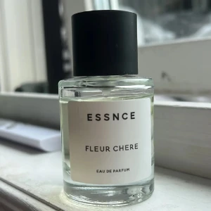 Essnce Fleur Chere Eau de Parfum - Minimalistisk parfymflaska från Essnce med doften Fleur Chere. Flaskan är cylindrisk i genomskinligt glas med svart matt kork och vit etikett med svart text. Innehållet är ljust och fräscht, perfekt för dig som gillar stilren design och subtil elegans.