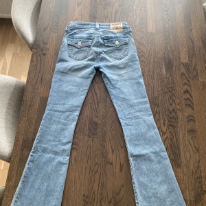 True Religion Becca Bootcut Jeans ljusblå - Säljer ett par ljusblå True Religion Becca Mid Rise Bootcut jeans i klassisk denim. Snygga detaljer på bakfickorna med knappar och kontrastsömmar. Bootcut-modell. Lite långa för mig som är 160 cm lång därför lite slitna nedtill. Skriv till mig om du vill ha fler bilder!