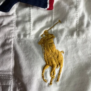 Vit långärmad piké från Ralph Lauren - Vit långärmad piké från Ralph Lauren med broderad gul polospelare, stort klubbmärke på bröstet och mörkblå siffran 3 på ärmen. Kragen är i marinblått och rött, vilket ger en sportig och klassisk look. Perfekt för dig som gillar preppy stil.