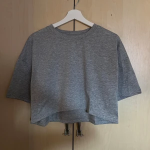 Grå croppad t-shirt i bomull - Enkel och stilren grå t-shirt med croppad passform och korta ärmar. Tillverkad i mjuk bomull, perfekt för en avslappnad look. T-shirten har rund halsringning och en något boxig siluett som ger en modern vibe.