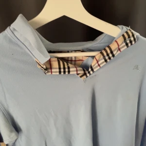 Ljusblå pikétröja från Burberry - Snygg ljusblå pikétröja från Burberry med klassisk rutdetalj i kragen och diskret broderad logga på bröstet. Tröjan har långa ärmar och är gjord i mjuk bomull, perfekt för en clean och stilren look.