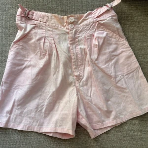 Ljusrosa shorts från Villardo - Ljusrosa shorts från Villardo i luftig bomull med hög midja. De har knäppning framtill med två knappar, bältesdetalj i midjan och resår baktill för extra komfort. Liten missfärgning högst upp men stör inte när de sitter på. Perfekta för varma dagar och en chill look.