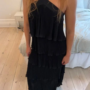 Svart långklänning med volanger - Supersnygg svart långklänning med flera lager volanger och breda axelband som korsas framtill. Klänningen har en plisserad struktur och är ärmlös, vilket ger en cool och elegant vibe. Från märket Design by Si, storlek XS/S