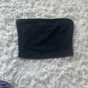 Svart bandeau tubtopp - Enkel och stilren svart bandeau tubtopp i stretchigt material. Toppen är axelbandslös och sitter tight runt kroppen, perfekt att matcha med höga jeans eller kjol. Snygg basic som funkar till många olika looks.