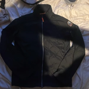 Svart Parajumpers jacka med orange foder - Säljer en svart Parajumpers jacka i storlek S med orange quiltat foder och klassisk PJS-patch på ärmen. Jackan har hel dragkedja framtill, hög krage och diskreta fickor med dragkedja. Perfekt för dig som vill ha en stilren och sportig look.