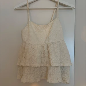 Beige volangtop från Gina Tricot - Supersöt beige linnetopp från Gina Tricot med tunna axelband och dubbla volanger. Toppen har en krinklad struktur som ger en cool och trendig look. Perfekt för varma dagar när du vill känna dig extra fin och avslappnad.