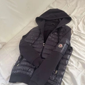 Monckler cardigan - Svart pufferjacka från Moncler med glansig quiltad front och ärmficka med logga. Jackan har huva, dragkedja och mjuka ärmar i bomullsmix. Perfekt för dig som vill ha en stilren och sportig look med premiumkänsla.