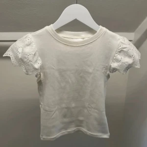 Vit blus med spetsärmar - Supersöt vit blus med korta puffärmar i broderad spets och rund halsringning. Perfekt för dig som gillar romantiska detaljer och vill ha en feminin look. Toppens kropp är i mjuk bomull och ärmarna har vacker volangkant.