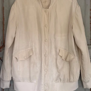 Beige bomberjacka med fickor - Other stories. Snygg beige lite oversized bomberjacka med klassisk passform och ribbade muddar. Jackan har två stora fickor med lock och tryckknappar framtill samt knäppning hela vägen. Perfekt för dig som gillar stilren och enkel design. Något noppig på muddarna.