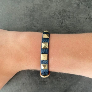 Valentino liknande armband  - Coolt armband i blått läder med fyrkantiga guldfärgade nitar längs hela bandet. Justerbart spänne i guldton och snygg struktur på lädret. Perfekt accessoar för att ge din outfit lite edge.