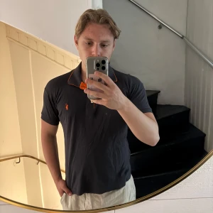 Svart pikétröja Polo Ralph Lauren M - Svart pikétröja från Polo Ralph Lauren med orange detaljer på kragen och logga på bröstet. Klassisk pikékrage och korta ärmar. Tillverkad i mjuk bomull, perfekt för en clean och sportig look. Sitter mer som S