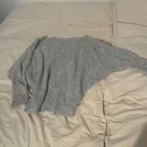 Grå stickad tröja off shoulder - Mysig grå stickad tröja med lång ärm och avslappnad passform. Tröjan har bred ribbad mudd vid ärmslut och nederkant, perfekt för lager på lager under kyliga dagar. Enkel och stilren design som funkar till det mesta.