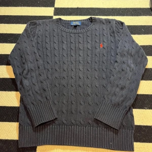 Polo Ralph Lauren tröja - Kabelstickad mörkblå tröja från Polo Ralph Lauren med klassisk rund hals och röd broderad logga på bröstet. Tröjan har ribbade muddar och nederkant, perfekt för dig som gillar en tidlös och clean stil.