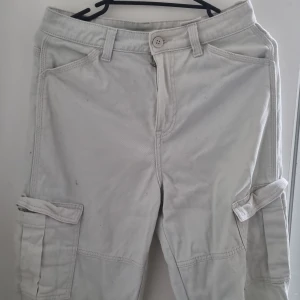 Beige cargobyxor med fickor - Säljer ett par ljusgrå cargobyxor med stora sidofickor och klassisk passform. Byxorna har raka ben, bälteshällor och är tillverkade i ett slitstarkt bomullstyg. Perfekta för en avslappnad och cool stil. Köpte byxan för länge sen typ 2 år sen men har använt dem tre gånger bara