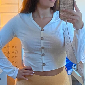 Vit croppad ribbad blus med knappar - Säljer en vit croppad blus med långa ärmar och snygga träknappar framtill. Toppen är ribbad och har v-ringning, vilket ger en fräsch och trendig look. 