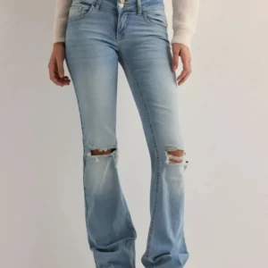 Ljusblå slitna bootcut jeans - Säljer ett par ljusblå jeans med bootcut-modell och slitna detaljer vid knäna. Jeansen har klassisk femficksdesign, normal midja och är tillverkade i mjukt denimtyg. Perfekta för en avslappnad och trendig look.