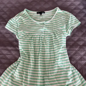 Grönvit randig t shirt - Söt t shirt i grönt och vitt med horisontella ränder. Klänningen har rund halsringning, tre knappar framtill och korta ärmar. Tillverkad i mjuk bomull, perfekt för varma dagar.