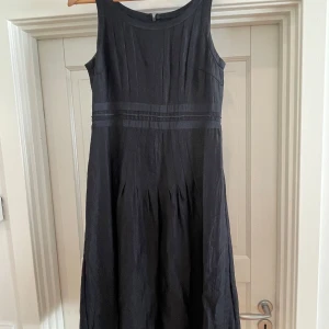 Svart långklänning i linne från Sandwich - Snygg svart långklänning från Sandwich i sidenlikt material, med ärmlös design och diskreta vertikala veck. Klänningen har markerad midja med banddetalj med små svarta paljetter och dragkedja bak. Perfekt för dig som gillar stilrena och klassiska plagg med en twist. Finns en kort kavaj i samma material som hör till. Båda delar ingår. 